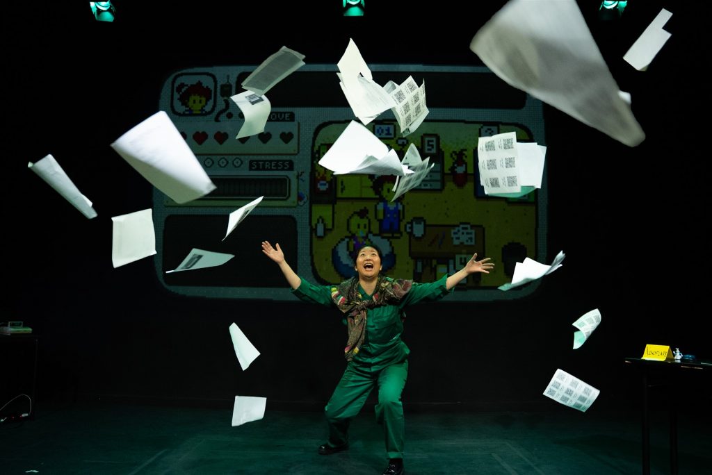 A performer stands center stage with their arms outstretched and an expression of excitement as dozens of sheets of paper fly dramatically through the air around them. They are dressed in a green outfit with a patterned scarf draped over their shoulders, crouching slightly beneath the swirling papers. The dark stage is illuminated by overhead spotlights, creating a strong contrast between the performer, the white pages, and the black background. Behind them, a large projected screen displays a colorful pixel-art video game interface. The game features a character inside a room with icons representing health and stress.